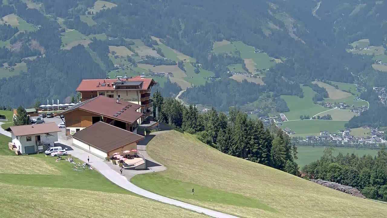A mountaintop restaurant complex overlooks a valley of green fields and a village under a sunny sky.