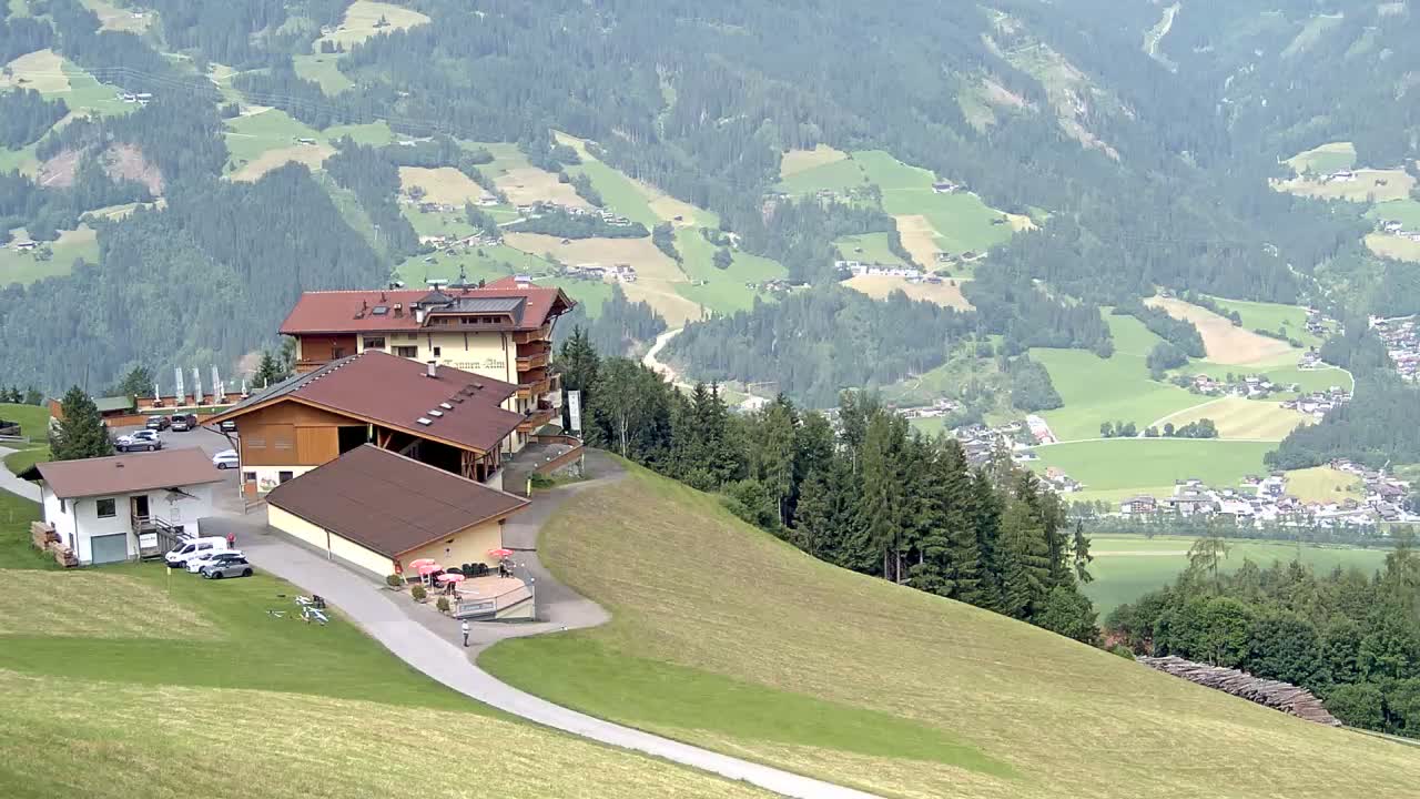A complex of buildings sits atop a grassy hill overlooking a valley of green fields and forests under a mostly clear sky.