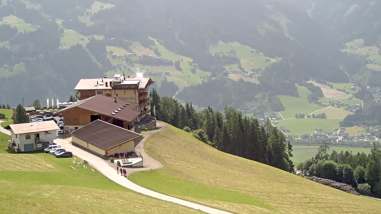 A mountaintop complex of buildings with a parking lot overlooks a hazy valley of green fields and distant villages under a sunny sky.