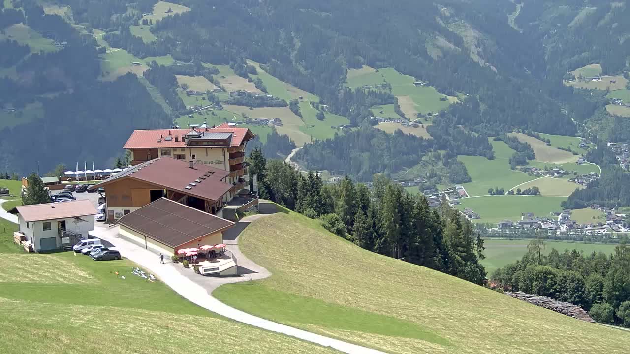A mountaintop complex of buildings with a parking lot overlooks a sunny valley of green fields and forests.