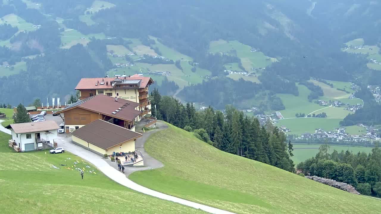 A mountaintop lodge with surrounding buildings and a paved path overlooks a valley of green fields and forests under an overcast sky.