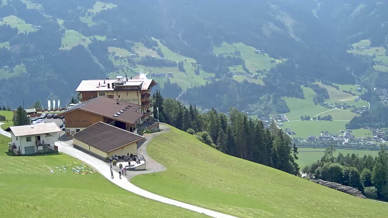 A mountaintop complex of buildings with a paved path leading down to a valley of green fields and a village in the distance under a clear, sunny sky.
