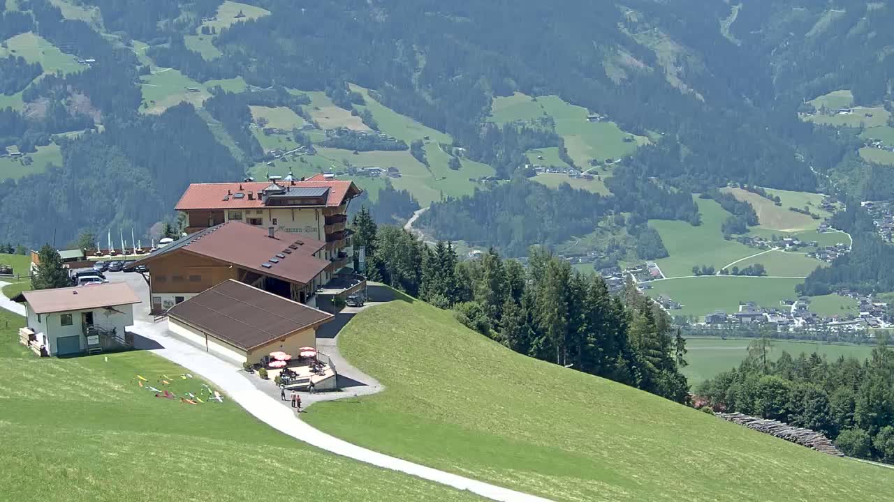 A complex of buildings sits atop a grassy hill overlooking a valley of green fields and forests on a sunny day.