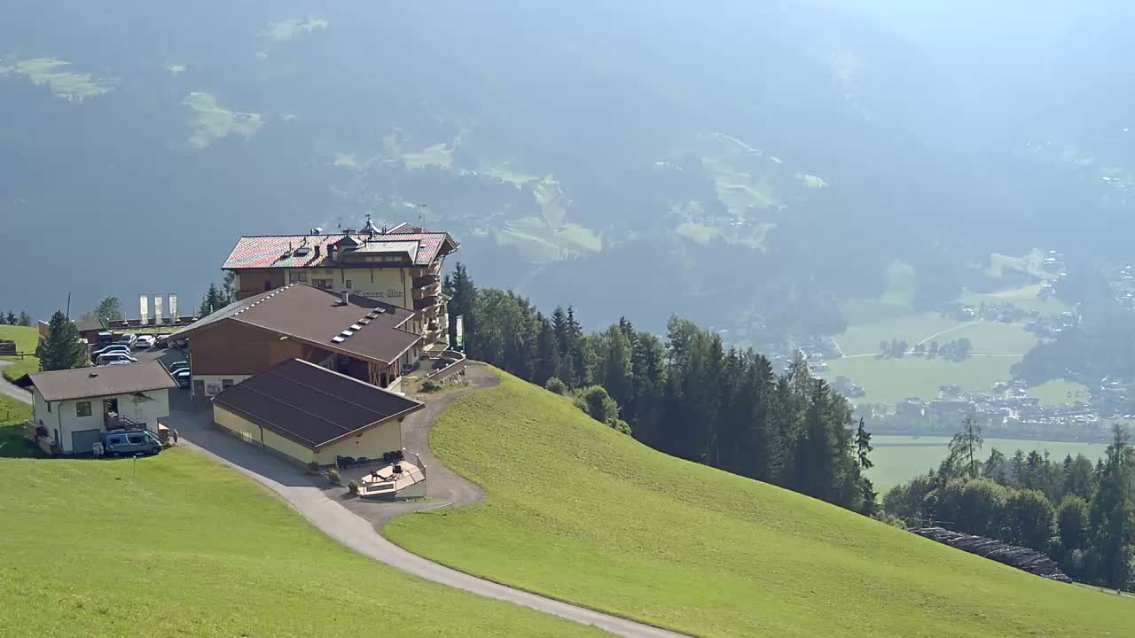 A complex of buildings sits on a grassy hillside overlooking a hazy valley and distant village under a bright, sunny sky.