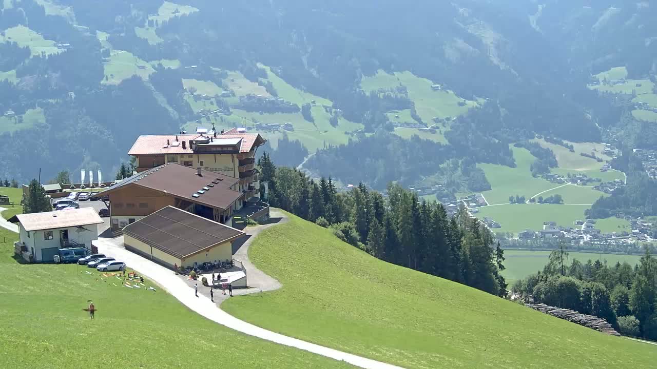 A mountaintop complex of buildings with a parking lot and a winding path overlooks a valley of green fields and distant villages under a clear, sunny sky.