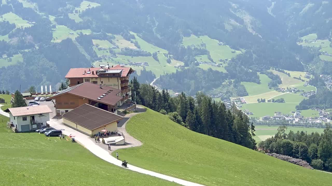 A mountaintop restaurant complex with a paved road leading down a grassy hillside overlooking a valley with fields and distant villages under sunny skies.
