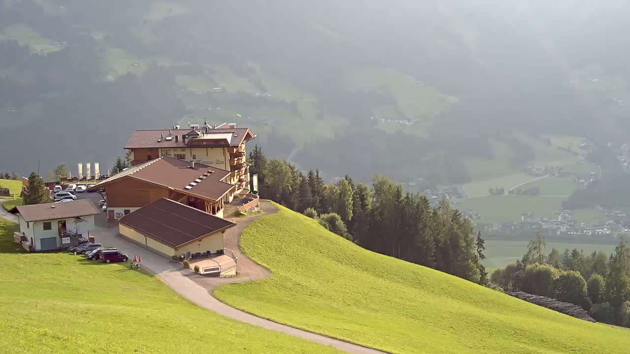 A multi-building complex sits atop a grassy hill overlooking a hazy valley and distant village.