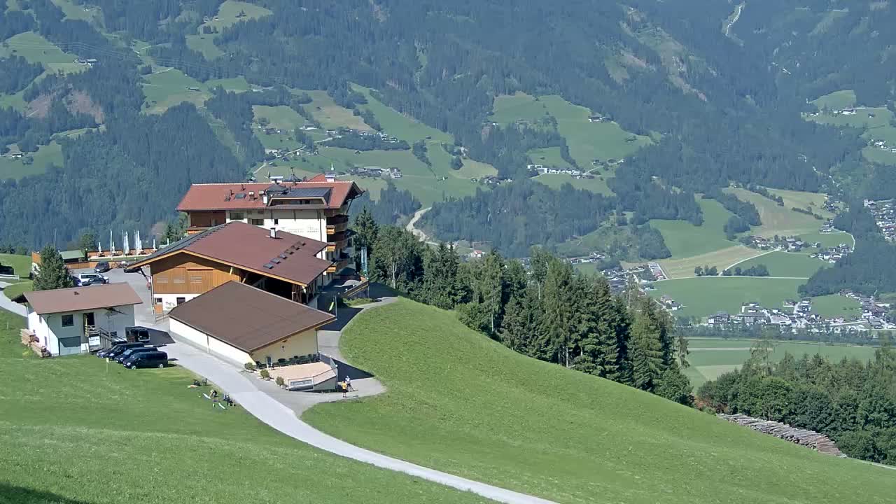 A cluster of buildings sits atop a grassy hillside overlooking a valley of green fields and forests under sunny skies.