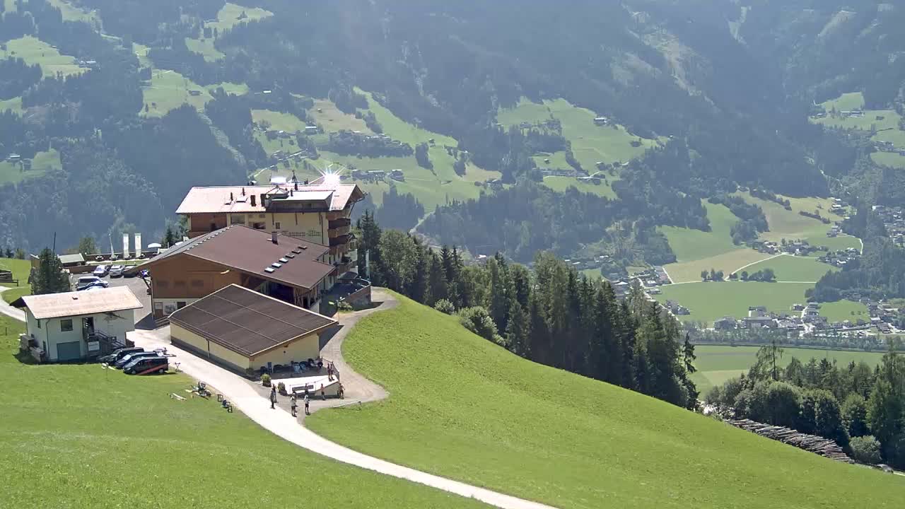 A mountaintop complex of buildings with a parking lot and a winding path overlooks a sunny valley dotted with houses and green fields.