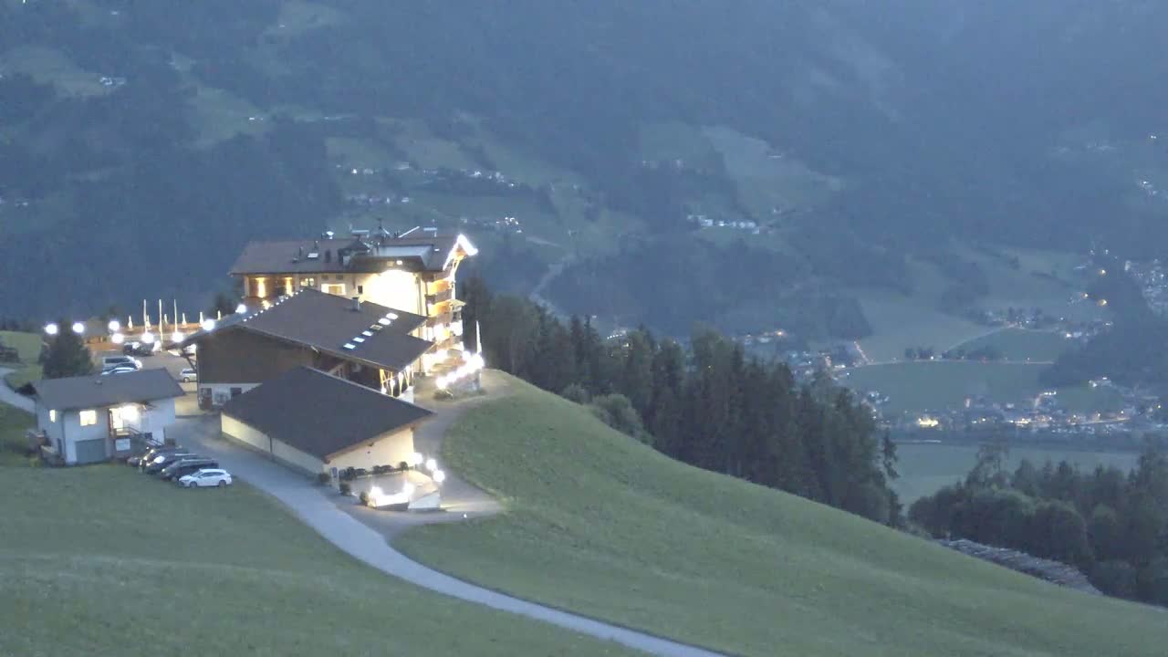 A brightly lit hotel complex sits atop a grassy hillside overlooking a valley at twilight.