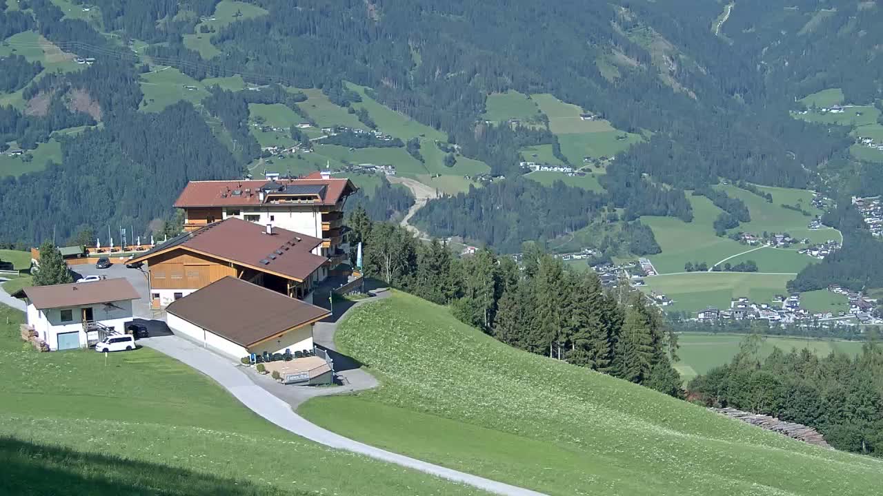 A mountaintop complex of buildings overlooks a valley of green fields and forests on a sunny day.
