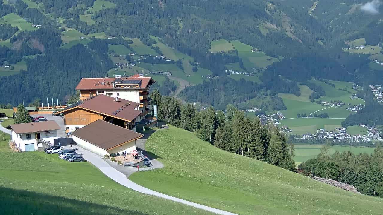 A hillside resort with several buildings and a parking lot overlooks a sprawling valley of green fields and forests under sunny skies.