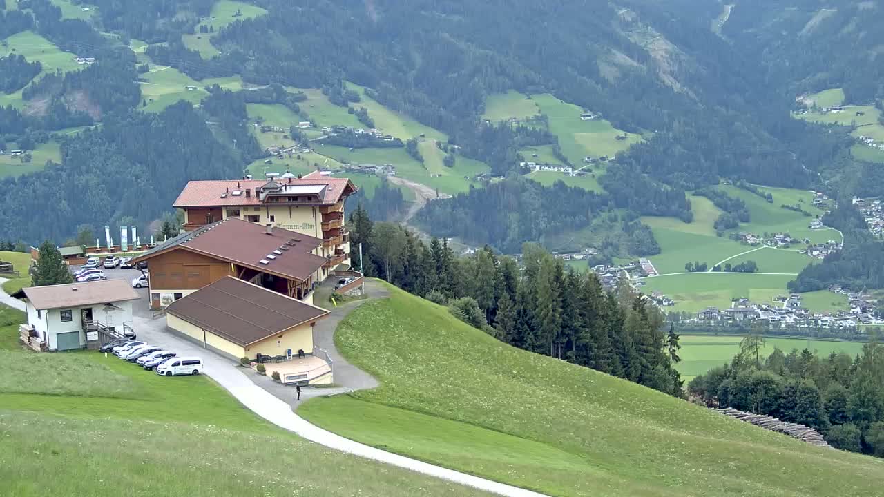 A hillside resort with several buildings and a parking lot overlooks a sprawling valley of green fields and forests under sunny skies.