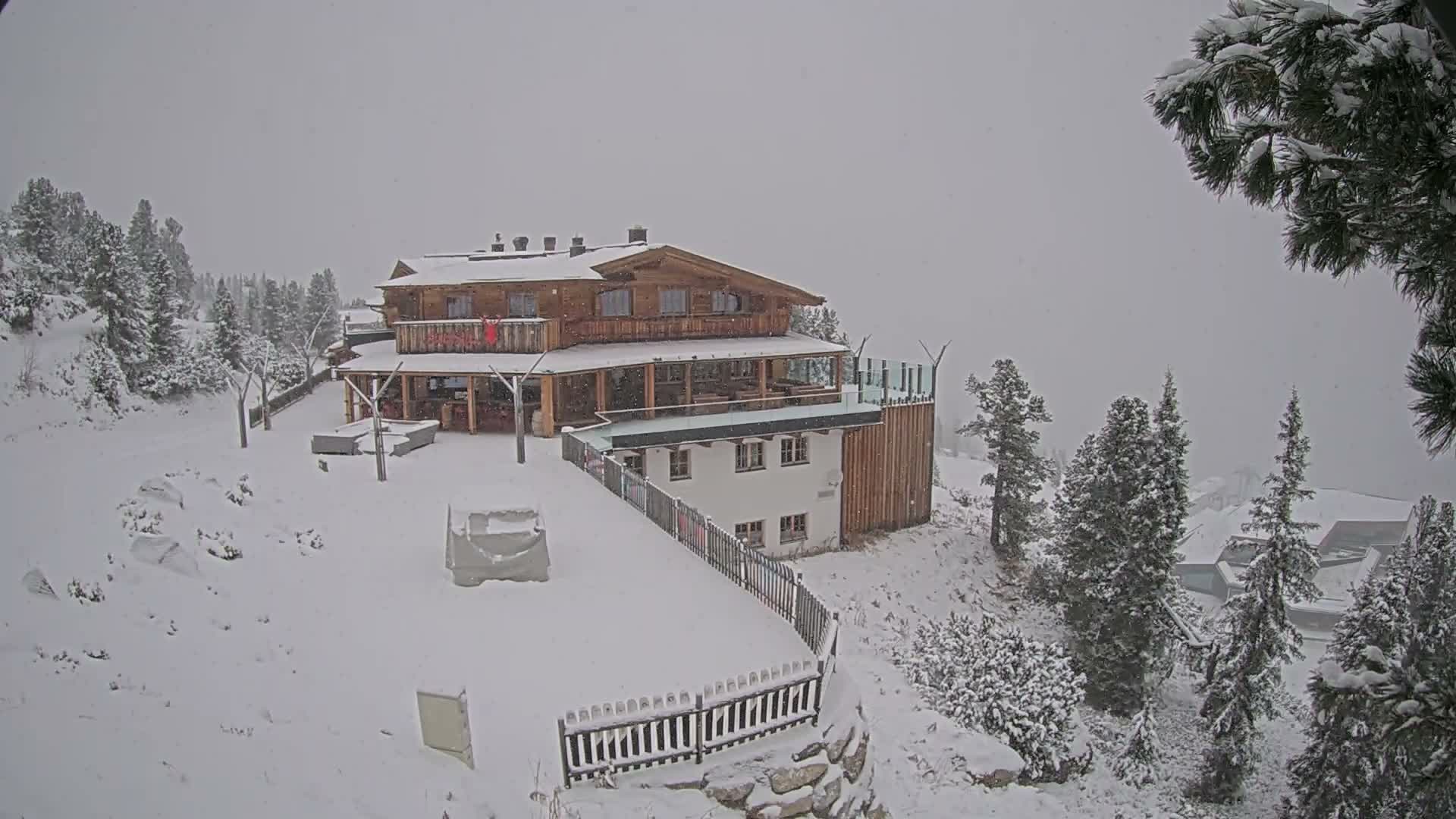 A large, multi-story wooden lodge and an attached white building are nestled on a snow-covered mountainside amidst numerous evergreen trees, under a gray, overcast sky with active snowfall.