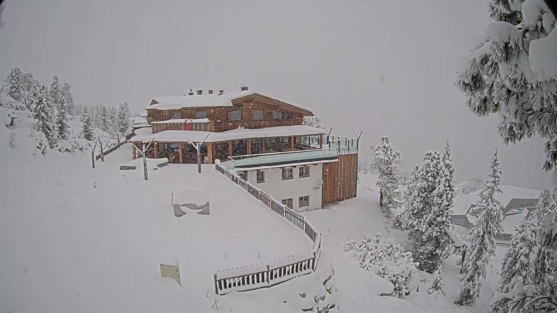 A sprawling wooden chalet with snow-covered roofs and multiple terraces, connected to a lower white building, is nestled on a heavily snow-laden mountain slope amidst numerous snow-covered pine trees, with active snowfall occurring under an overcast sky.
