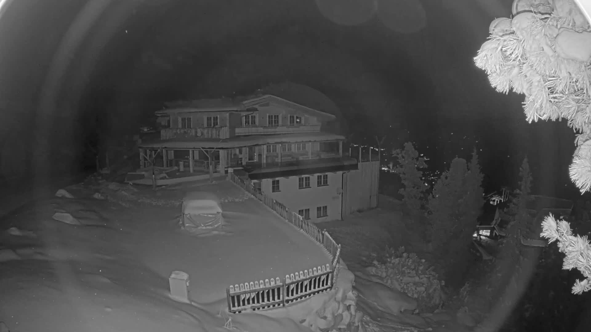 A clear, cold, and snowy night scene displays a multi-story alpine lodge and its snow-covered grounds, featuring a parked car and a fence, with distant town lights visible in the valley below and frosted tree branches partially framing the foreground.