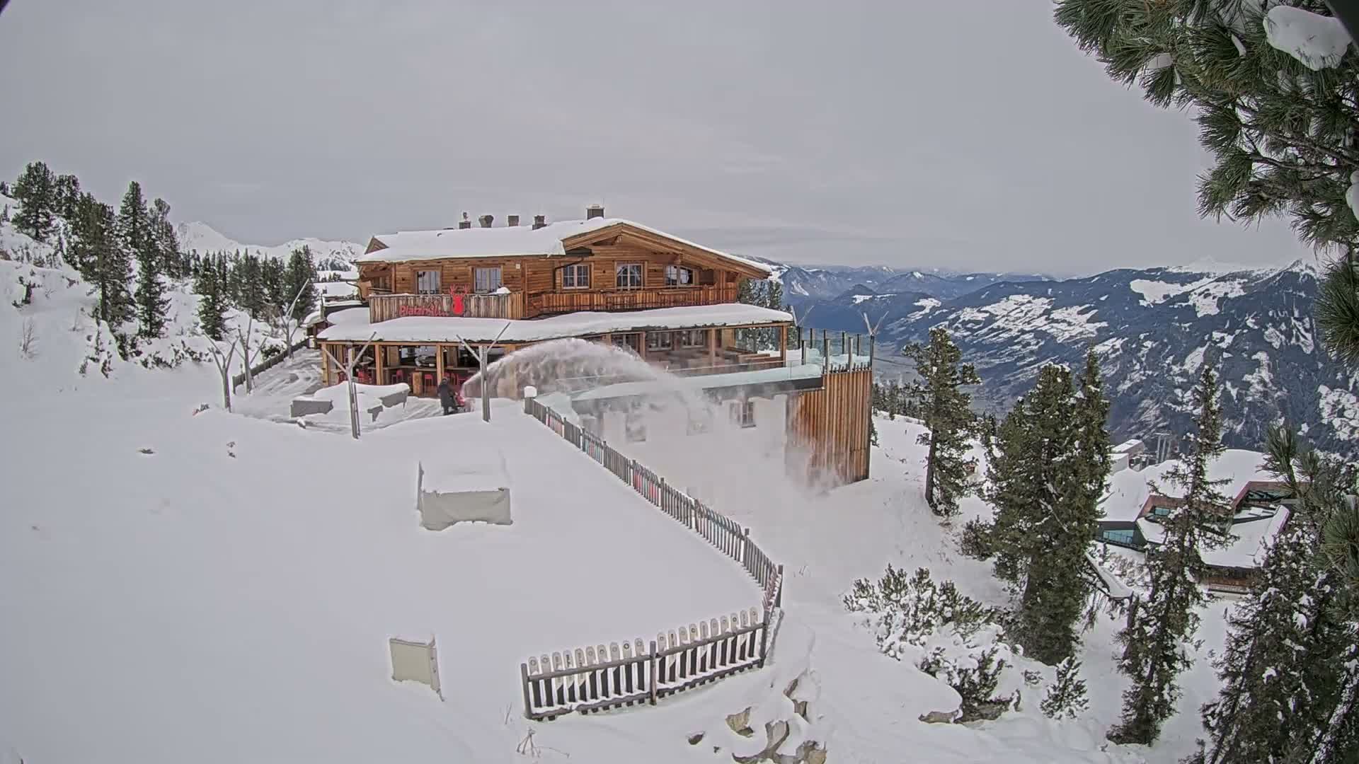 A snowblower vigorously clears a deep accumulation of snow from a path in front of a large, multi-level wooden mountain lodge, offering panoramic views of snow-covered peaks and valleys under an overcast sky.