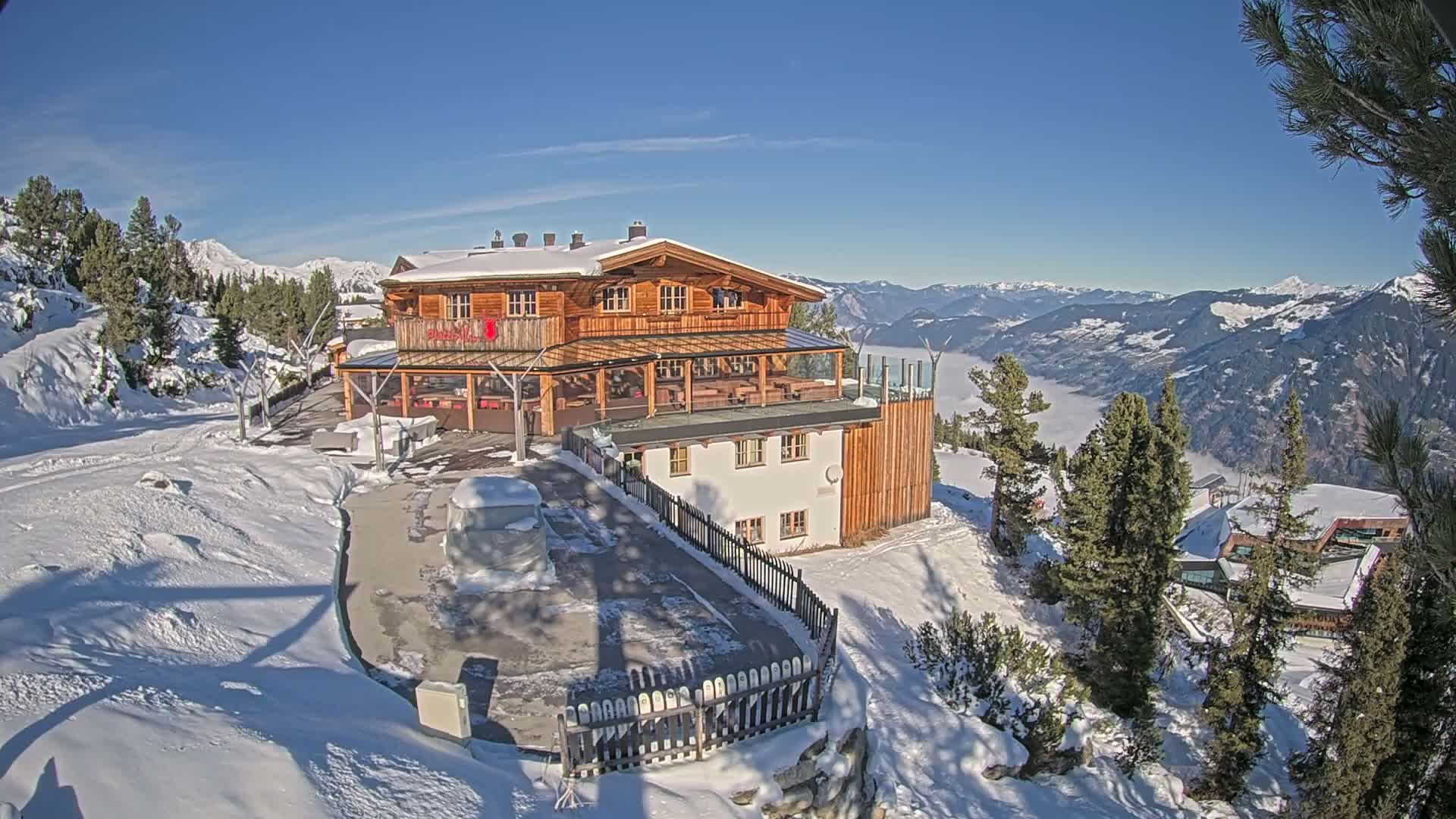 A multi-story mountain lodge, primarily wooden with a white lower level, sits on a snow-covered slope, providing a stunning vista of distant snow-capped mountains and a cloud-filled valley under a clear, sunny blue sky.