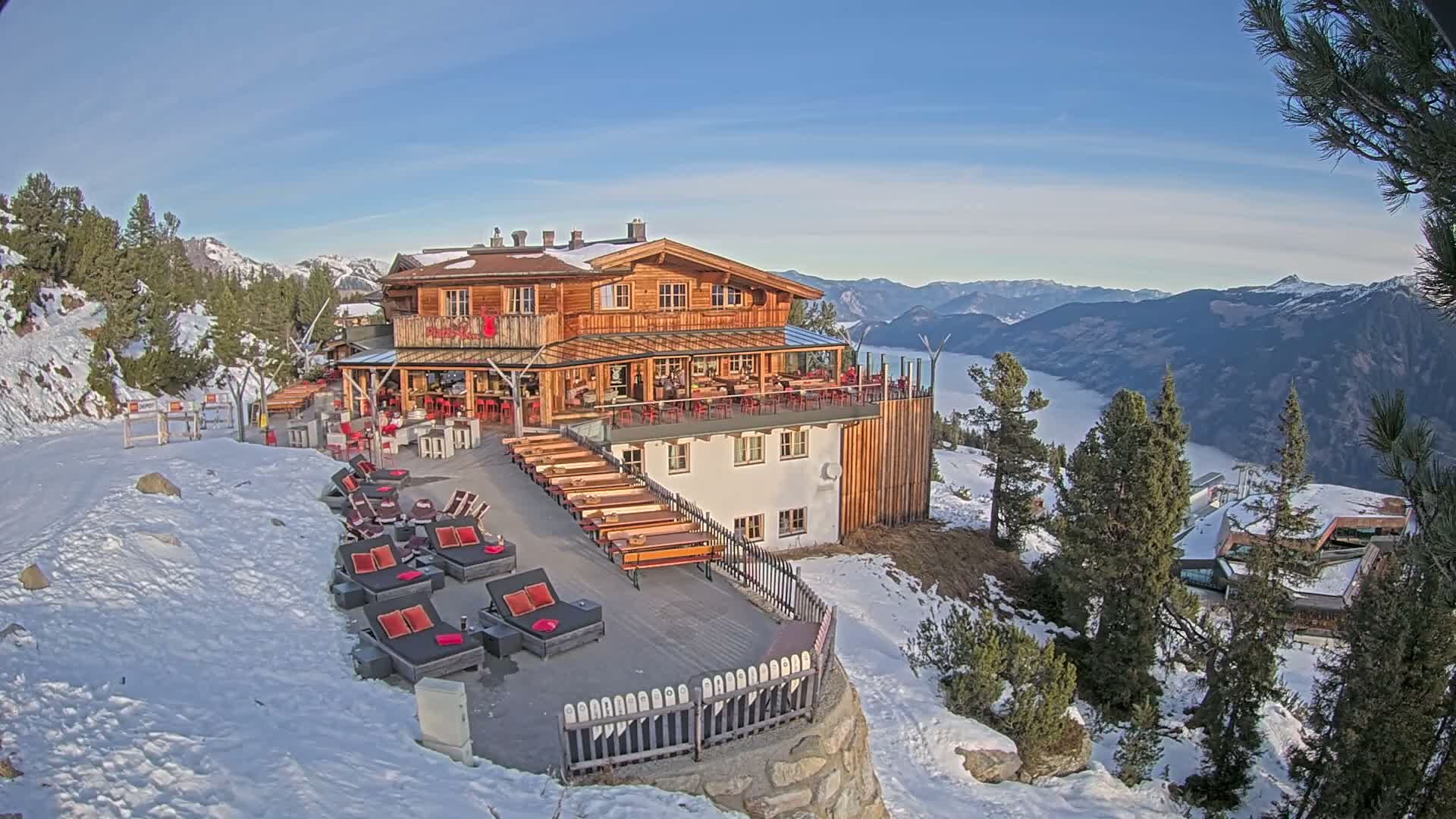 A rustic multi-story wooden mountain lodge with extensive outdoor seating and terraces is nestled on a snowy slope under a clear blue sky, offering panoramic views of distant peaks and a cloud-filled valley on a bright, sunny winter day.