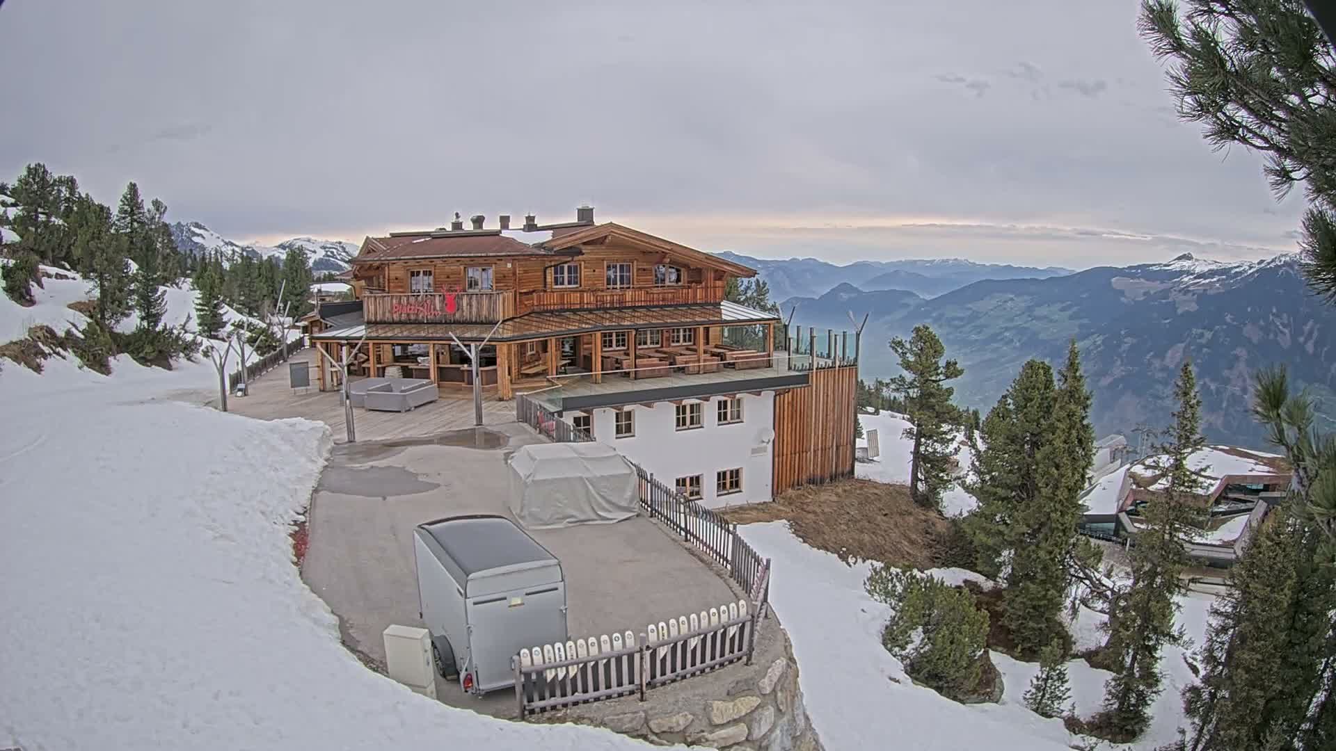 A rustic multi-story wooden mountain lodge with extensive outdoor seating and terraces is nestled on a snowy slope under a clear blue sky, offering panoramic views of distant peaks and a cloud-filled valley on a bright, sunny winter day.