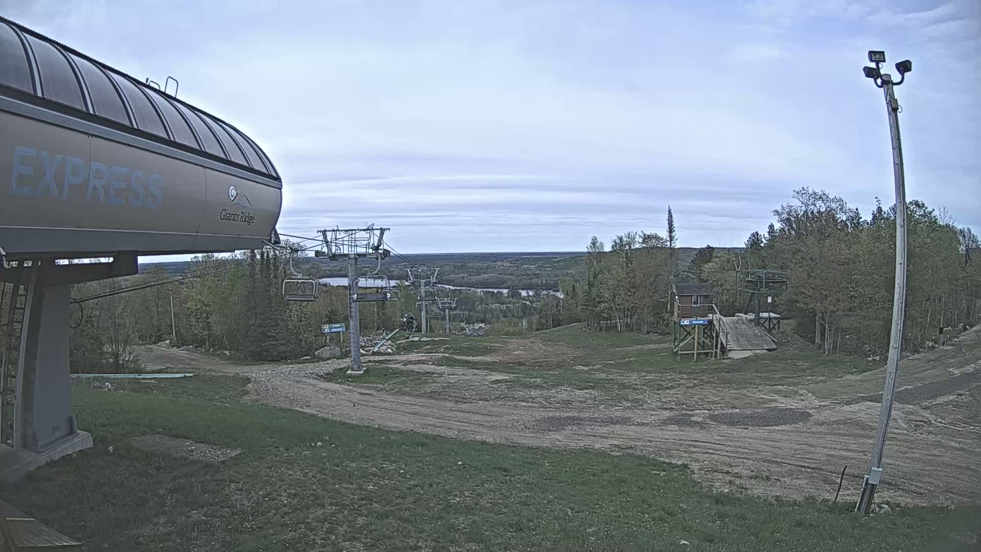 A ski lift base station with a chairlift ascending a hill towards a distant, tree-lined river valley under a partly cloudy sky.