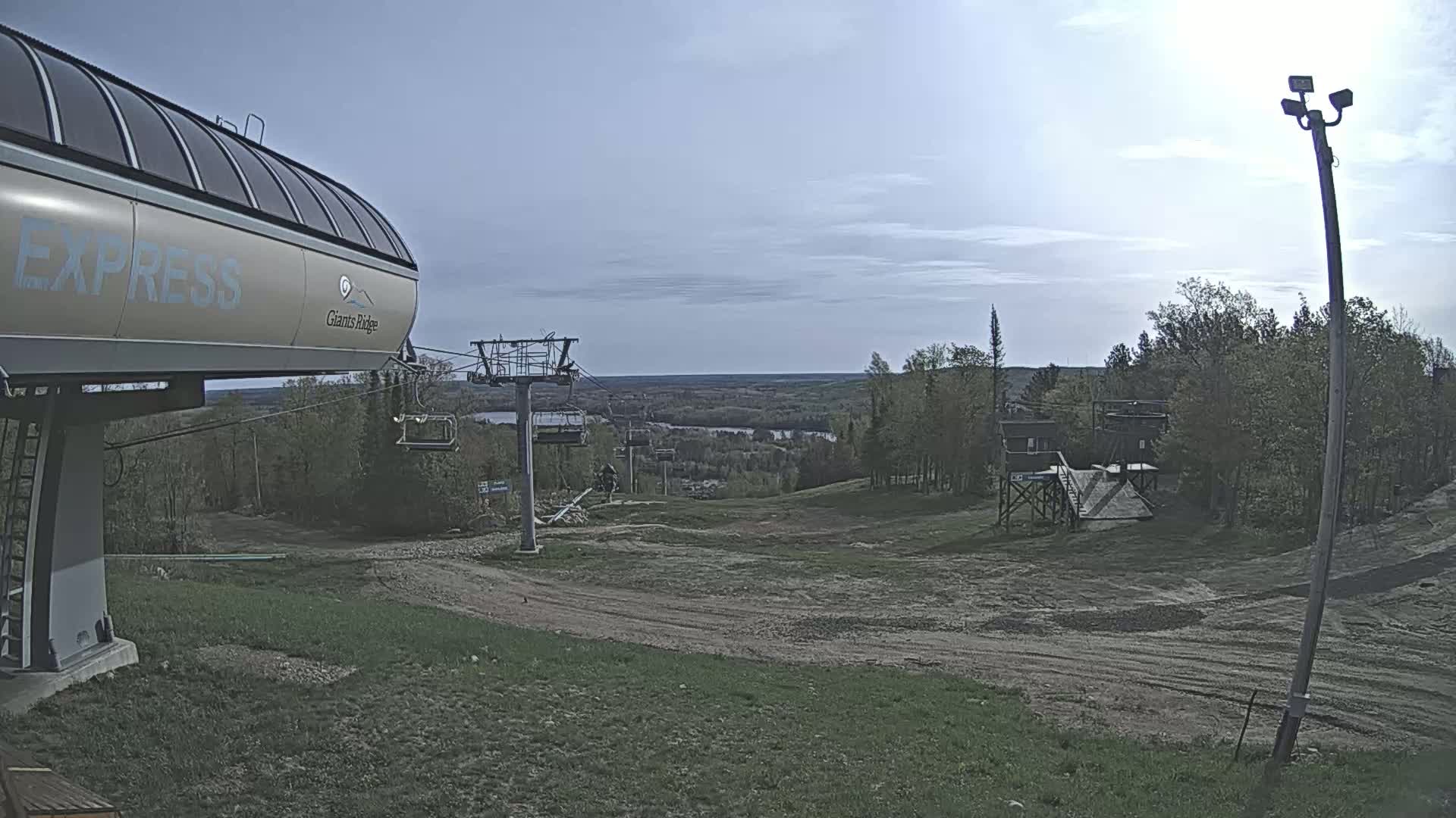 A chairlift and its base station are situated on a grassy hill overlooking a valley under a mostly clear sky.