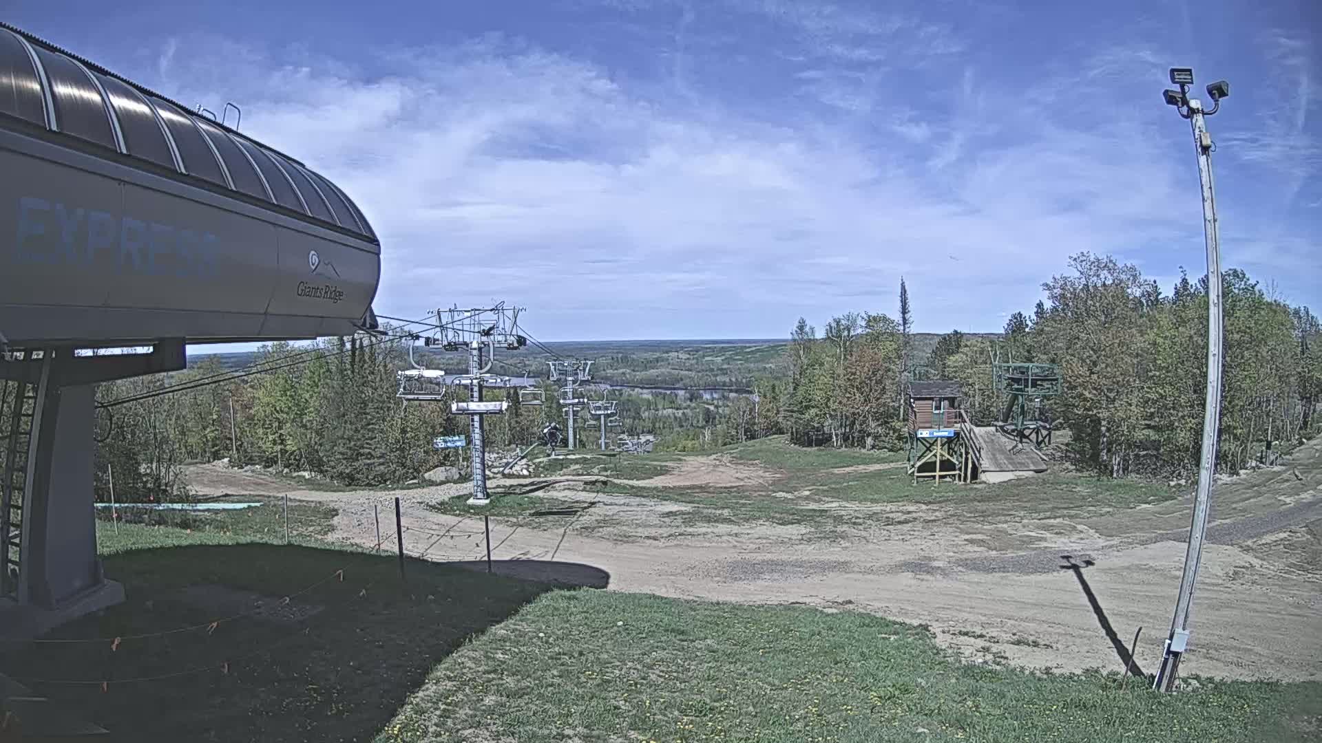 A ski lift base area with a chairlift and a mostly clear, sunny sky overlooks a treeline and distant flat land.