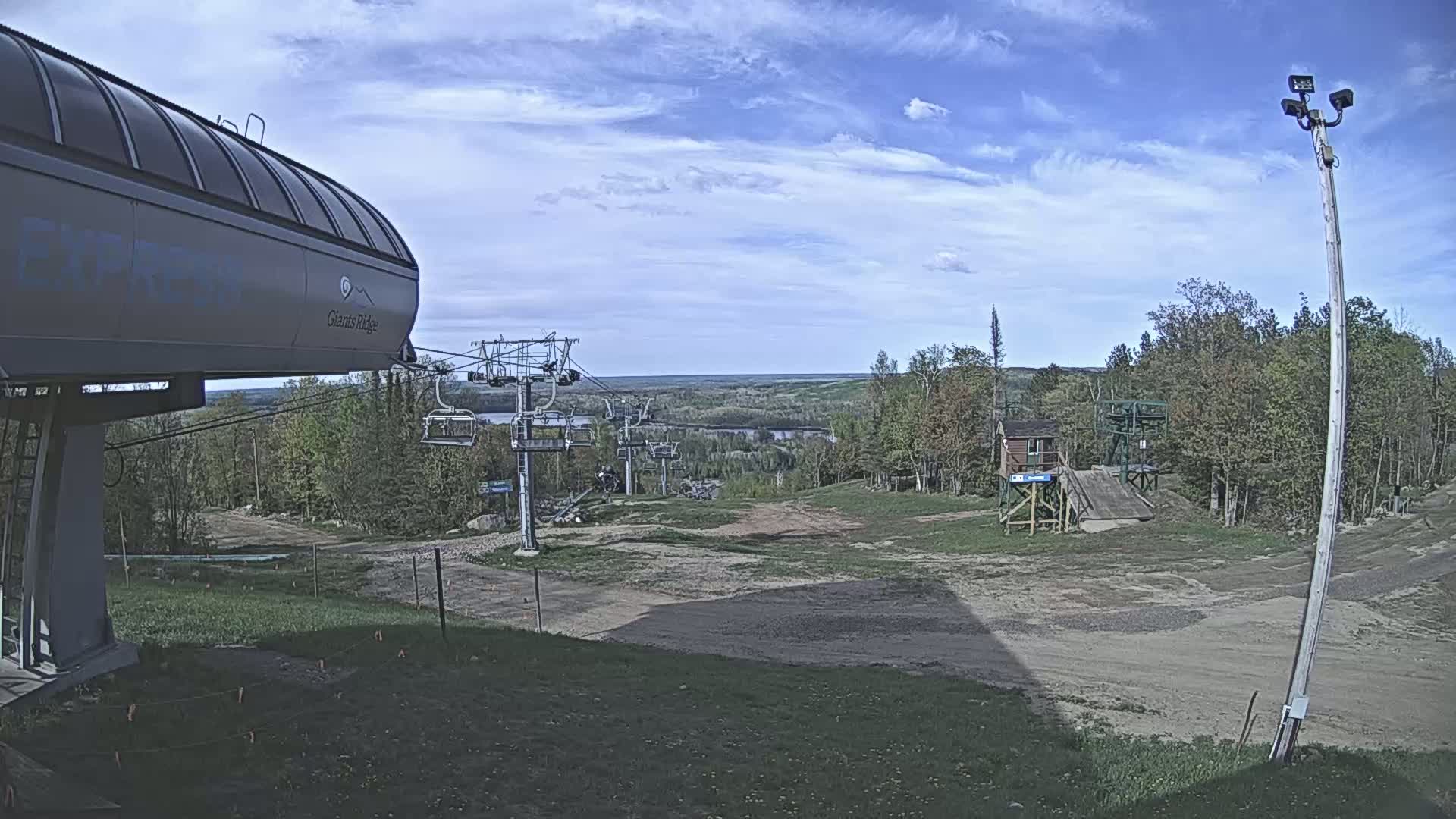 A ski lift with a chairlift visible in the distance under a partly cloudy sky, overlooking a grassy landscape and a river valley.