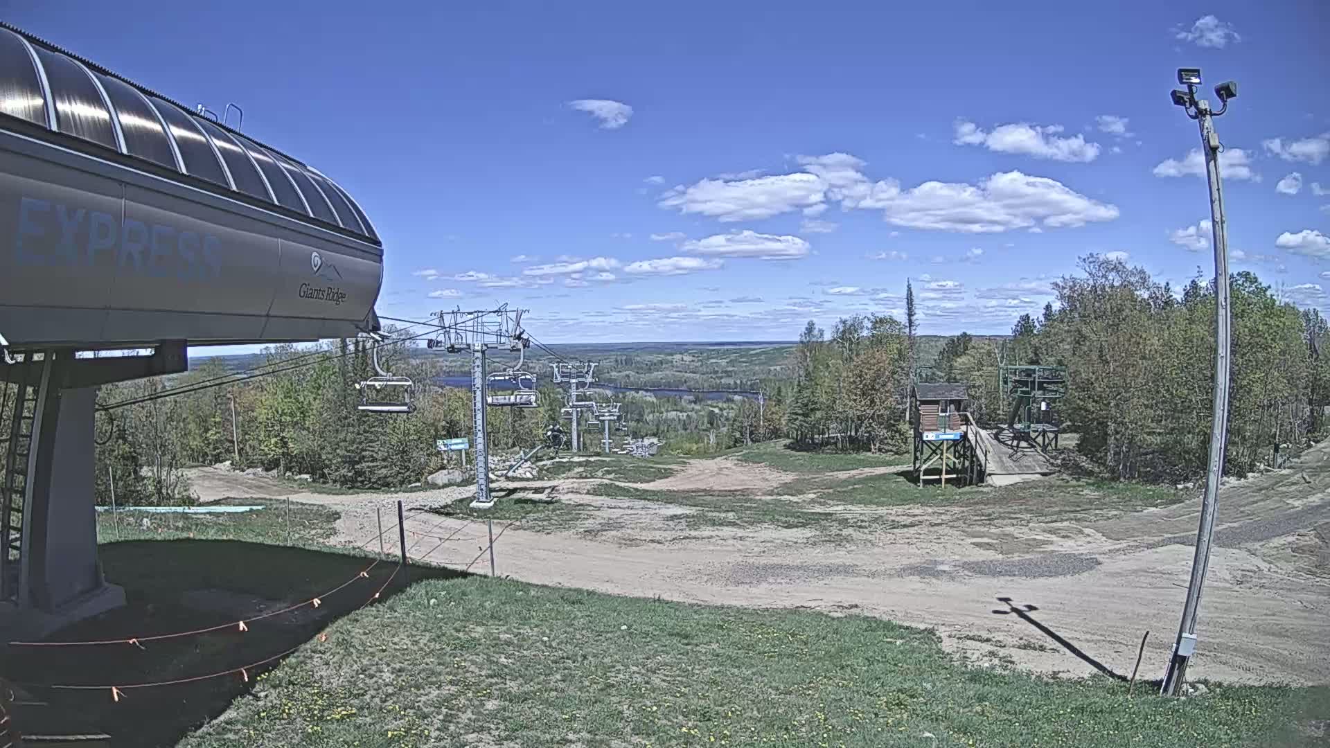 A ski lift base station with a chairlift extending up a hill toward a distant view of a lake and forest under a partly cloudy blue sky.