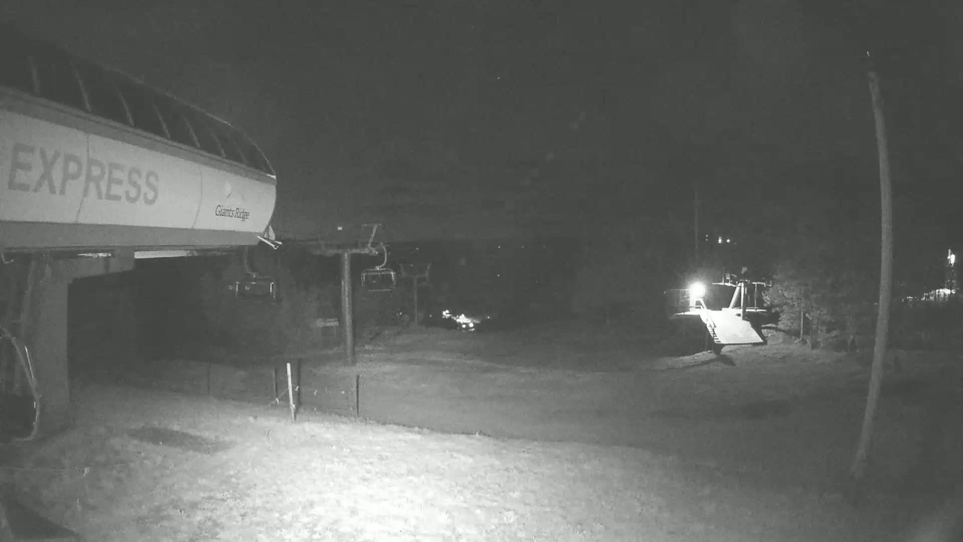 A nighttime, snow-covered scene shows a portion of a ski lift's base station with a chairlift in the background under dark, possibly cloudy conditions.