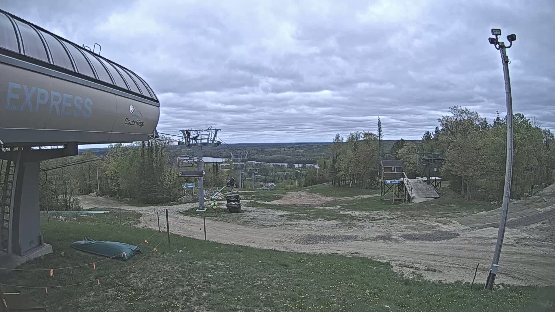 A ski lift base area with overcast skies shows a chairlift, a small building, and a mostly barren landscape with some green vegetation.
