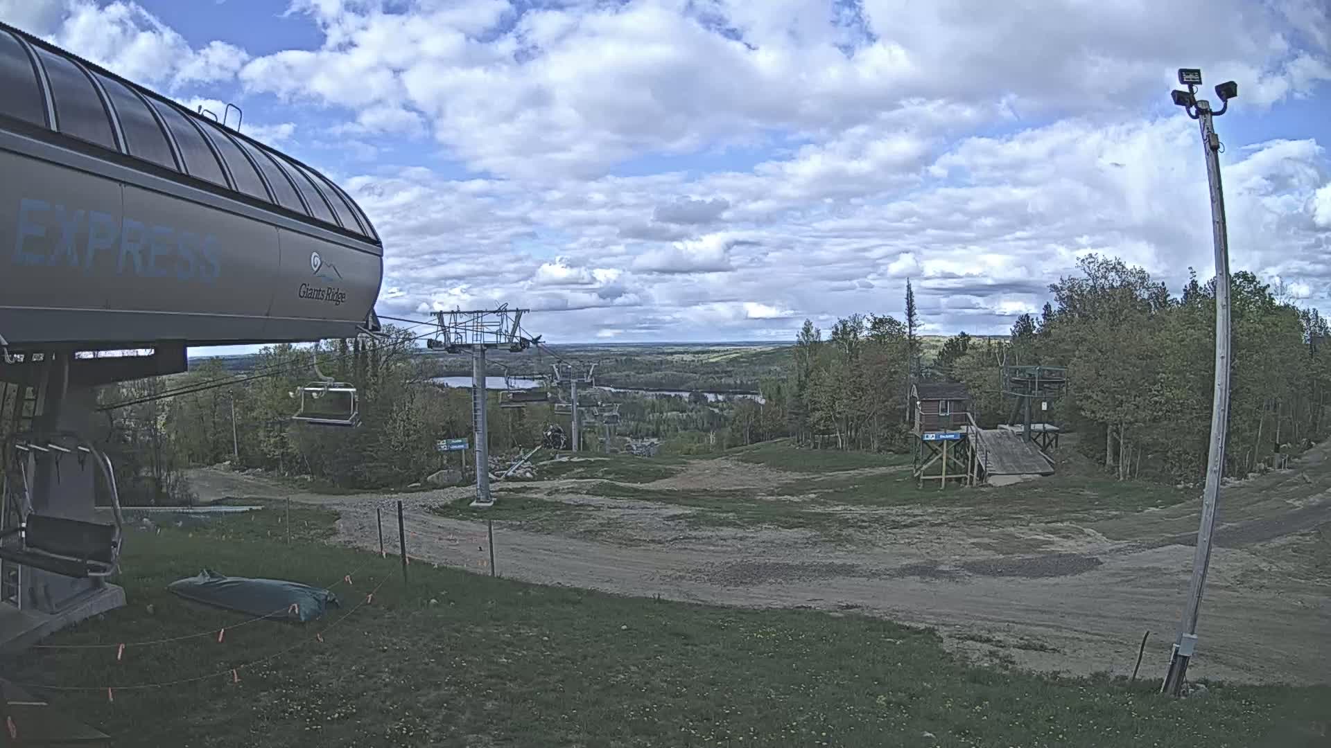 A ski lift base area with overcast skies shows a chairlift, a small building, and a mostly barren landscape with some green vegetation.