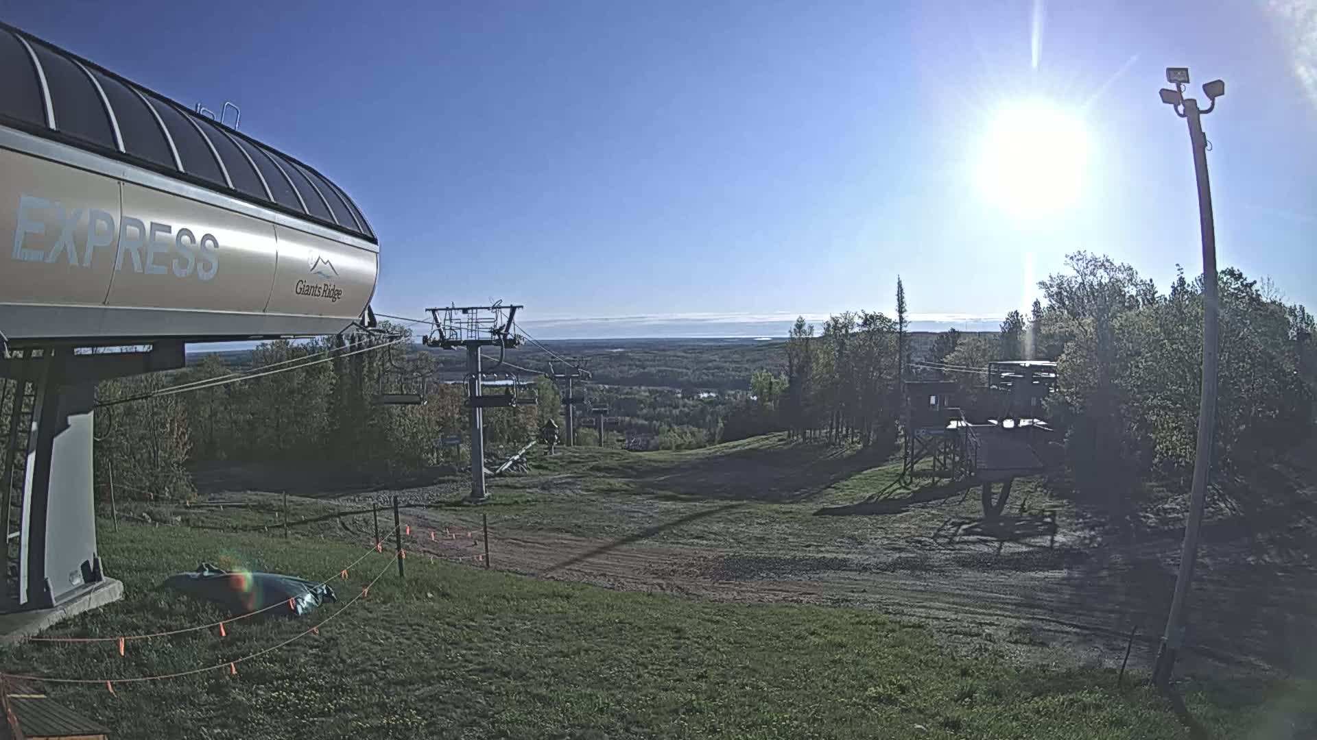 A ski lift base station sits on a grassy hill overlooking a wooded valley and a distant body of water under a bright, sunny sky.