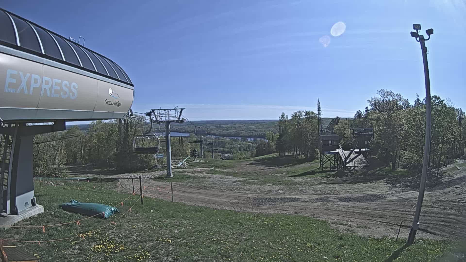 A ski lift terminal sits atop a grassy hill overlooking a valley and a lake under a clear, sunny sky.