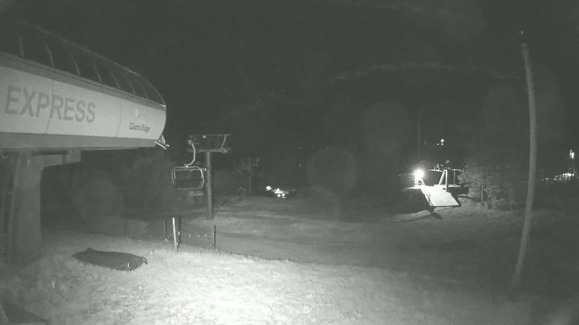 A nighttime, snow-covered ski lift area shows a chairlift and a snow-grooming machine under low-light conditions.