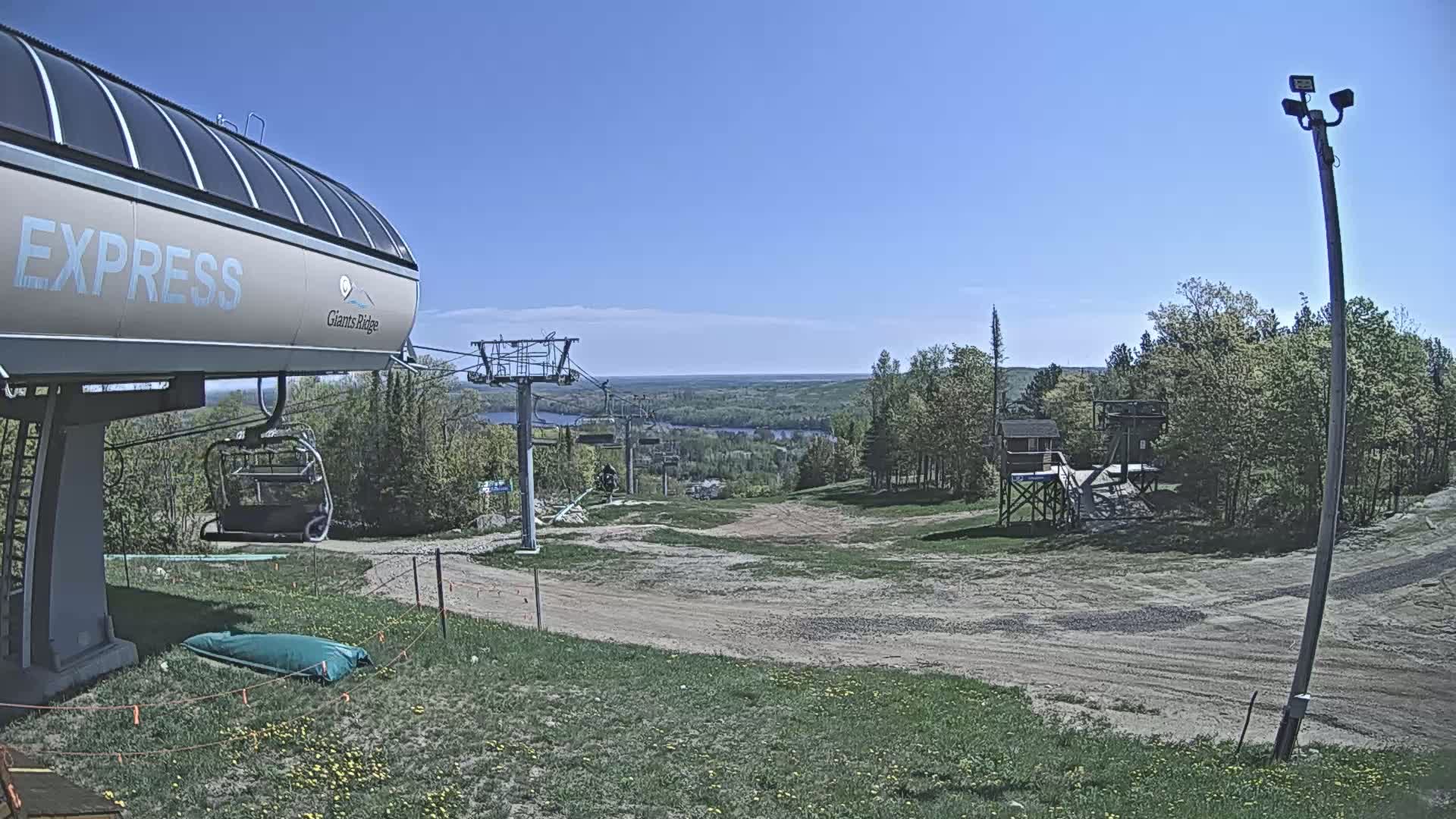 A ski lift base station sits on a grassy hill overlooking a mostly barren ski slope and a distant body of water under a clear blue sky.