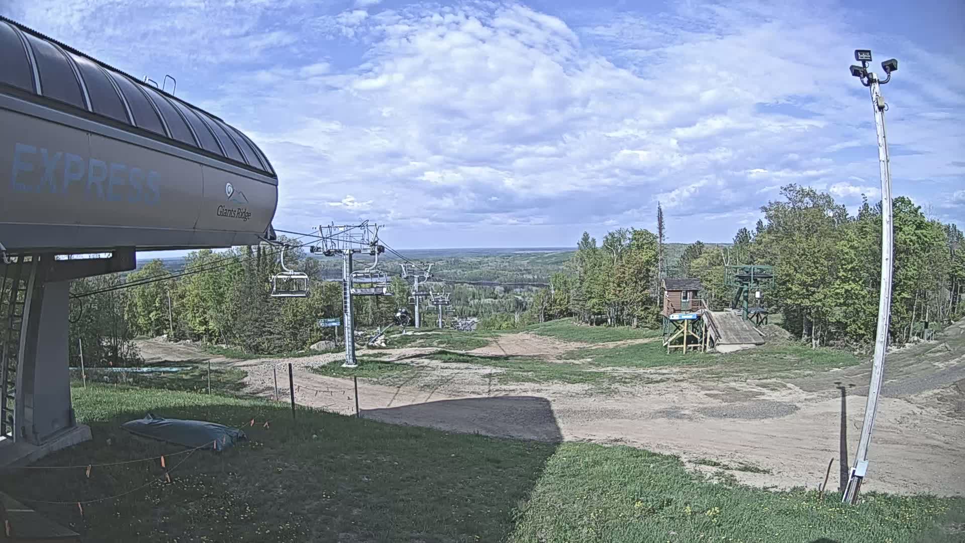 A ski lift base station with a chairlift extending uphill into a wooded area under a partly cloudy sky.