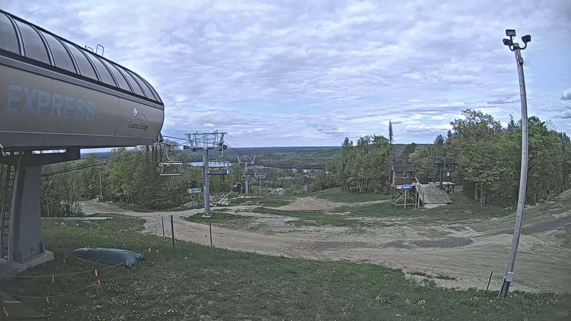 A ski lift base station with a chairlift extending into a wooded area under a partly cloudy sky.