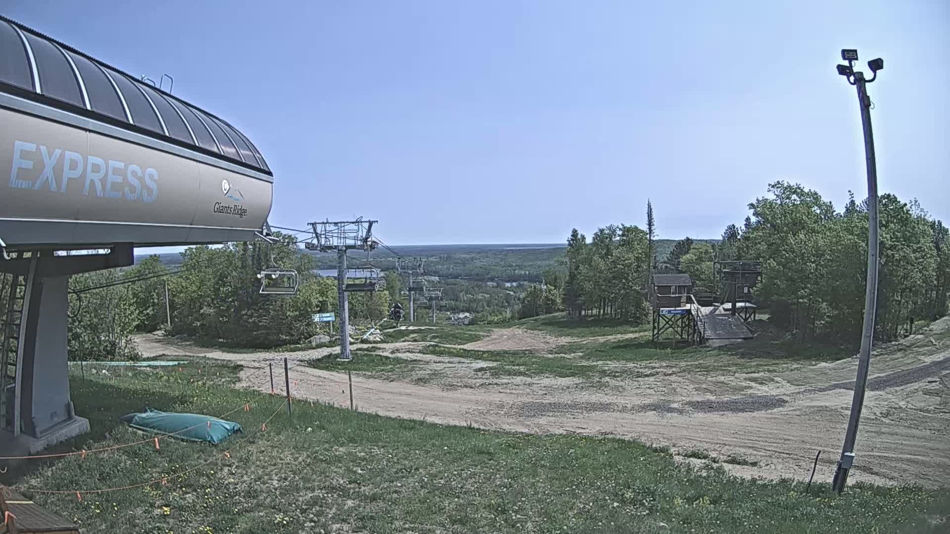A chairlift base station sits on a grassy hill overlooking a dirt path leading to a ski lift and a wooded area under a clear blue sky.
