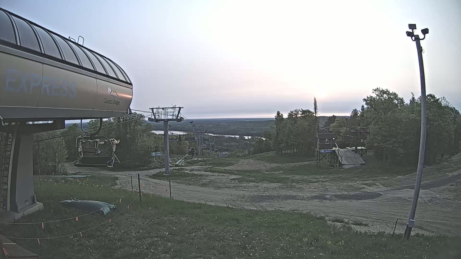 A ski lift terminal at sunset overlooks a grassy hill, a chairlift, and a distant body of water under a clear sky.