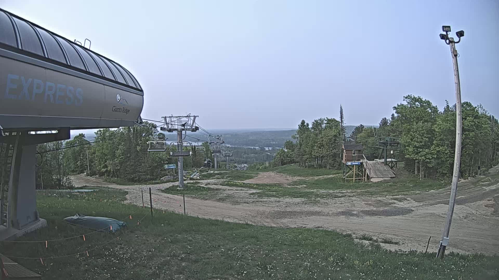 A ski lift base station with a chairlift extending into a treeline and a distant view of a valley under a clear, light-grey sky.