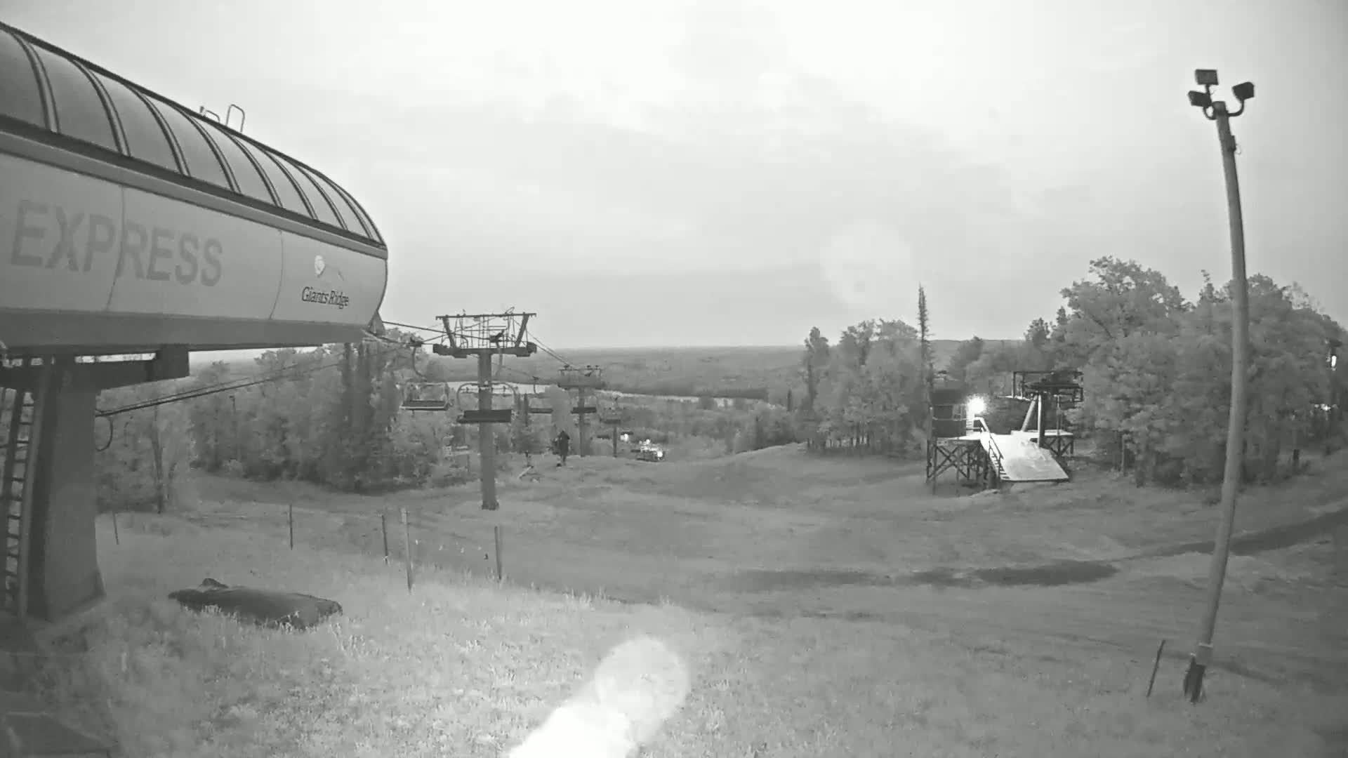 A ski lift base station with a chairlift extending uphill into a treeline under an overcast sky.