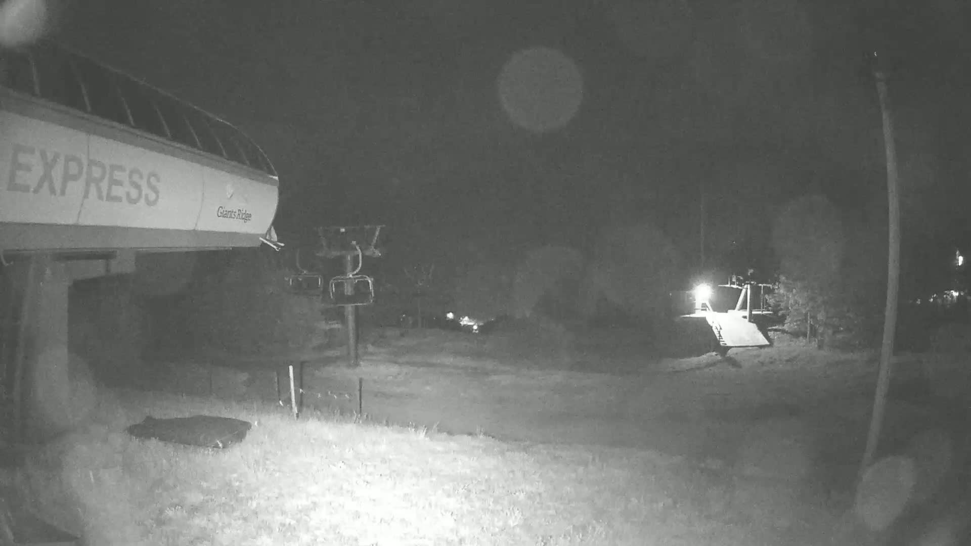 A nighttime, low-light image shows a chairlift and its base station at a ski resort, with some trees and grassy areas visible in the dark.