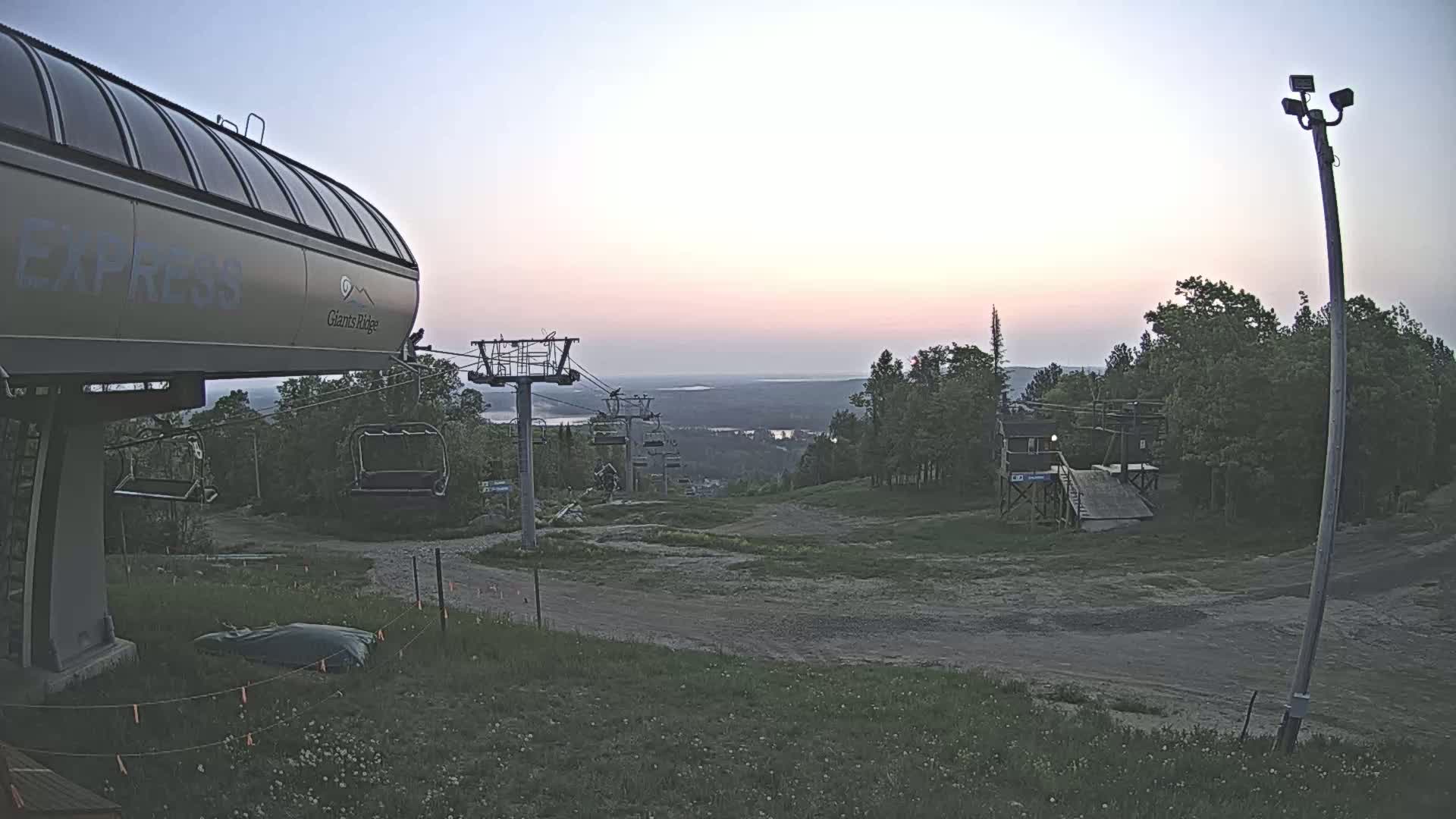 A ski lift base station at sunset overlooks a grassy hill and a distant body of water under a clear sky.
