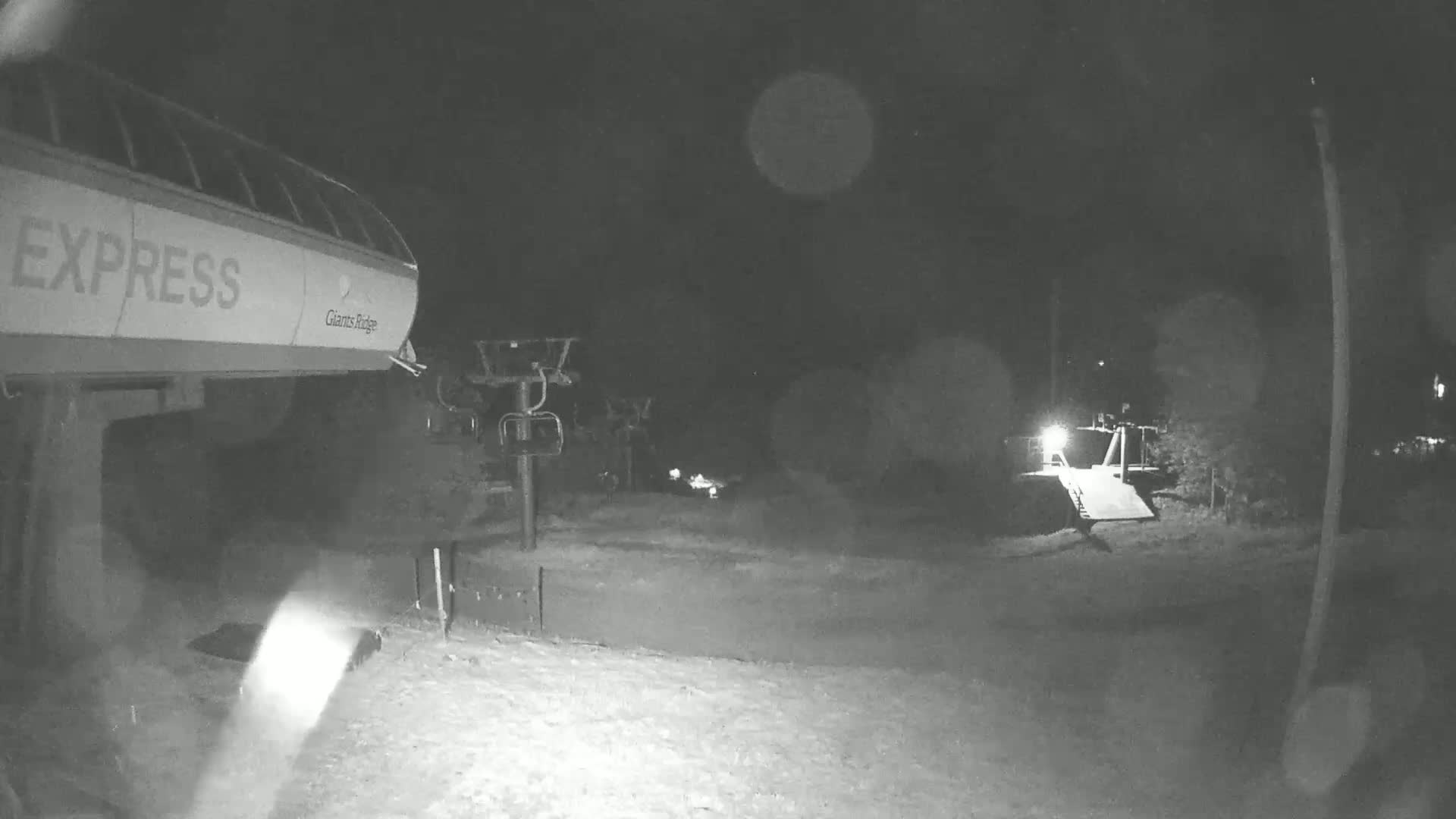 A nighttime, snow-covered ski lift base with a chairlift and a partially visible building.