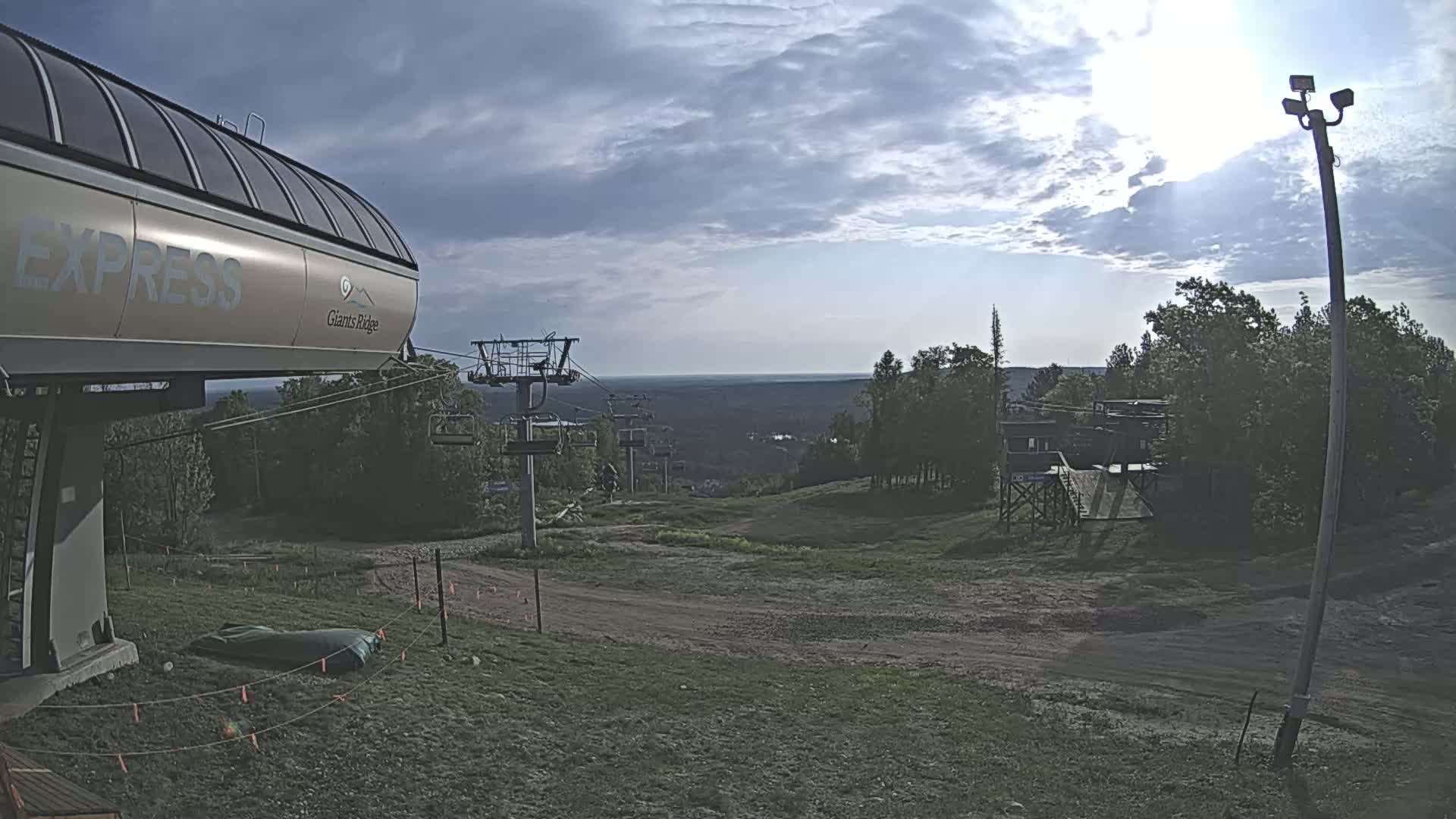 A ski lift base station sits at the top of a grassy hill under a partly cloudy sky, with a distant view of more hills and trees.