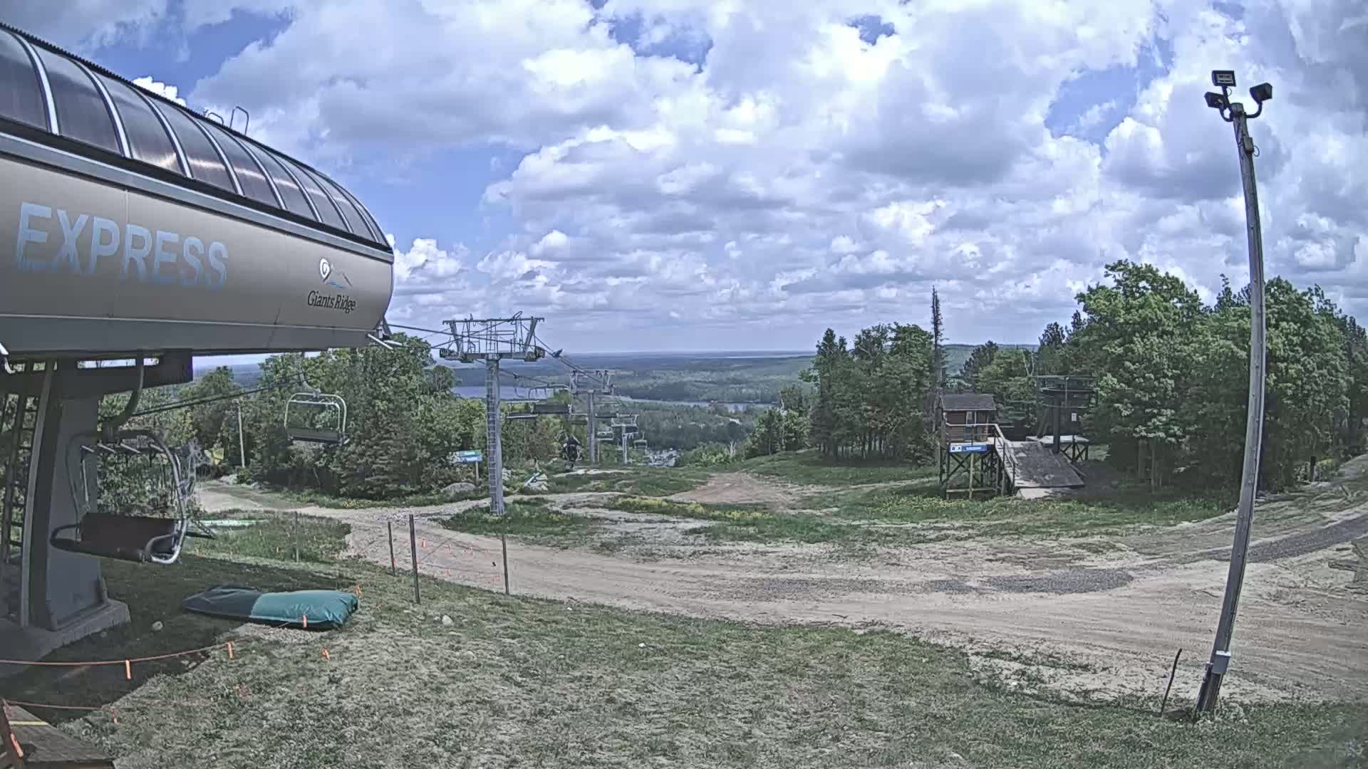 A chairlift, ski lift infrastructure, and a mostly dirt path are visible under a partly cloudy sky, overlooking a lake and forest in the distance.