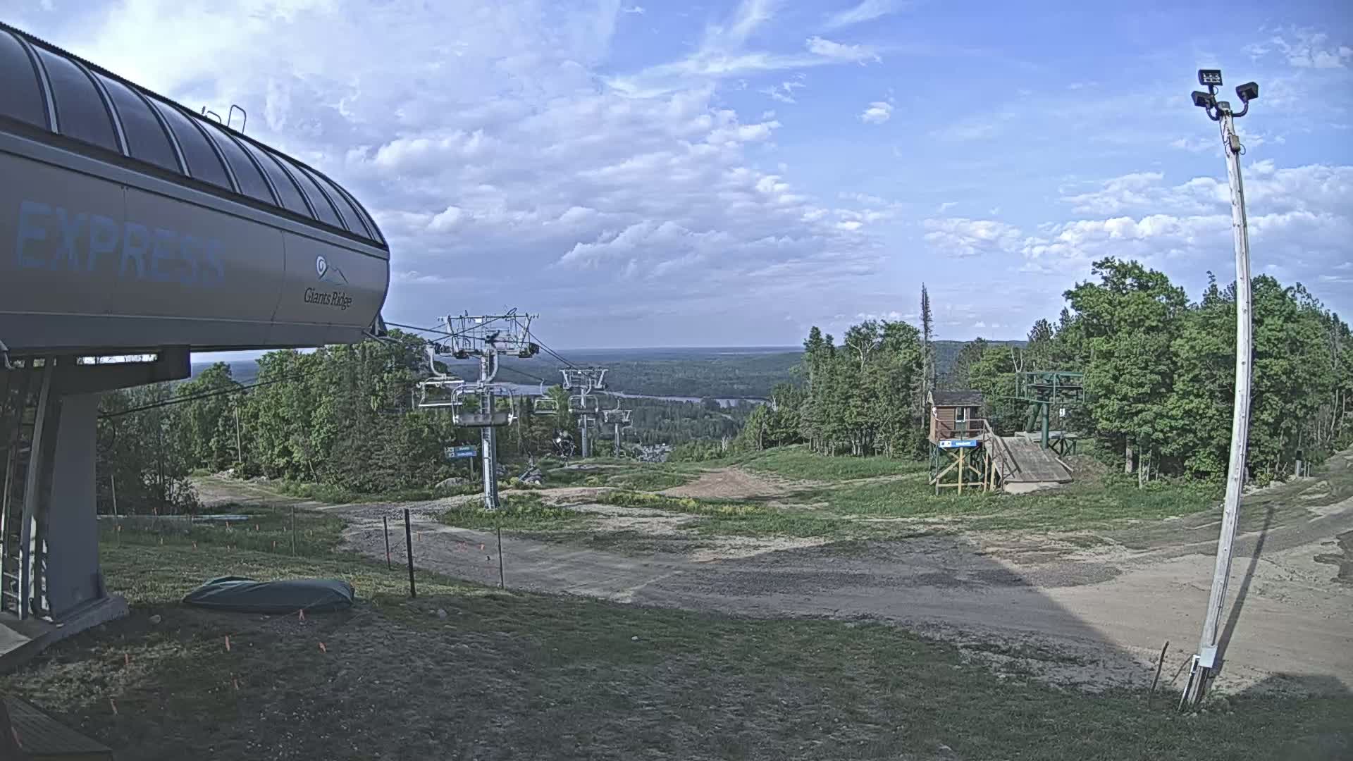 A mostly sunny view of a ski resort in summer shows a chairlift and a dirt path leading to a wooded area and a body of water in the distance.