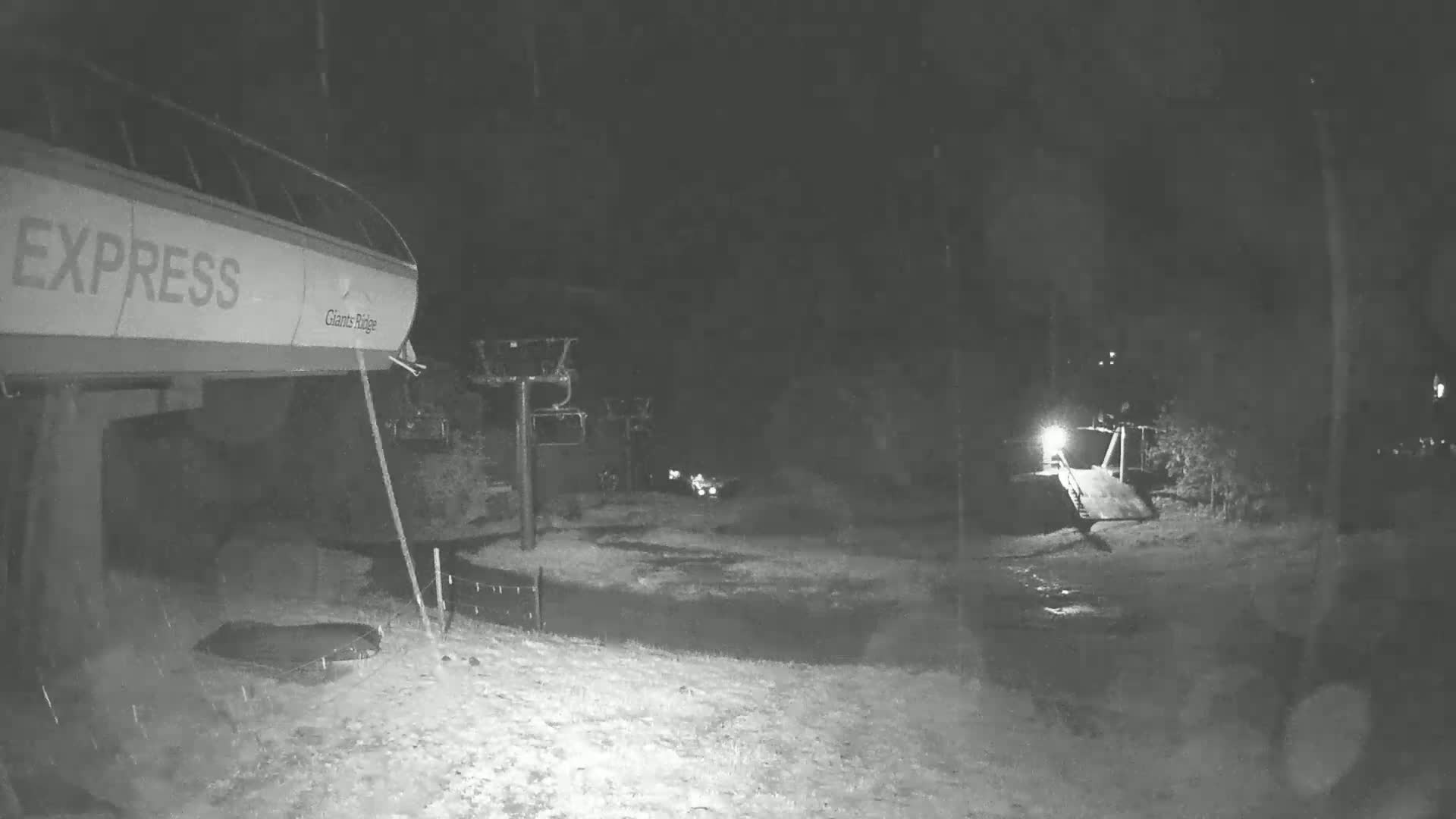 A nighttime, snowy scene shows a ski lift terminal and chairlift, with a slightly illuminated path leading away.