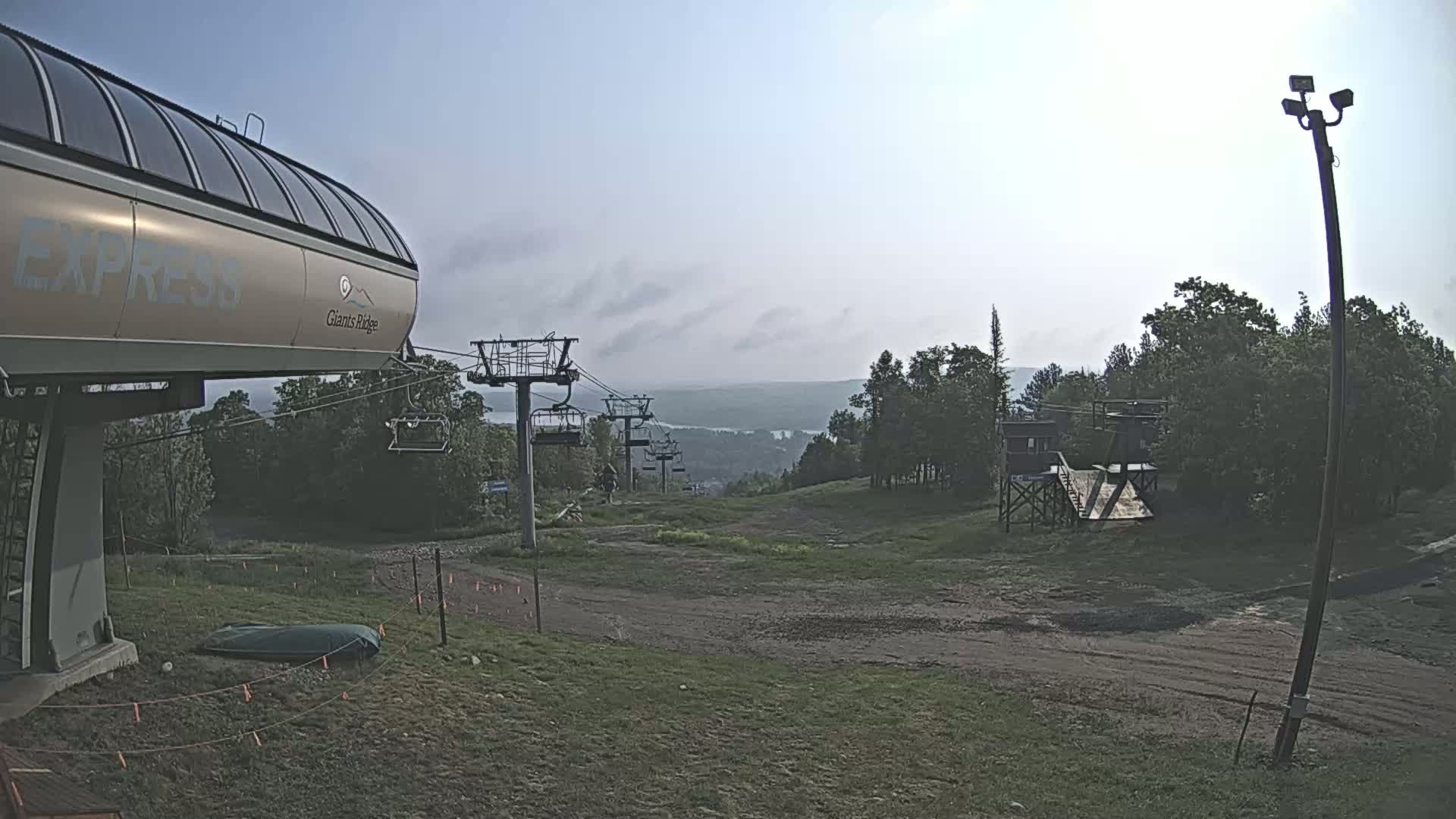 A chairlift base station and an empty ski lift extend towards a hazy, distant lake under a mostly clear sky.